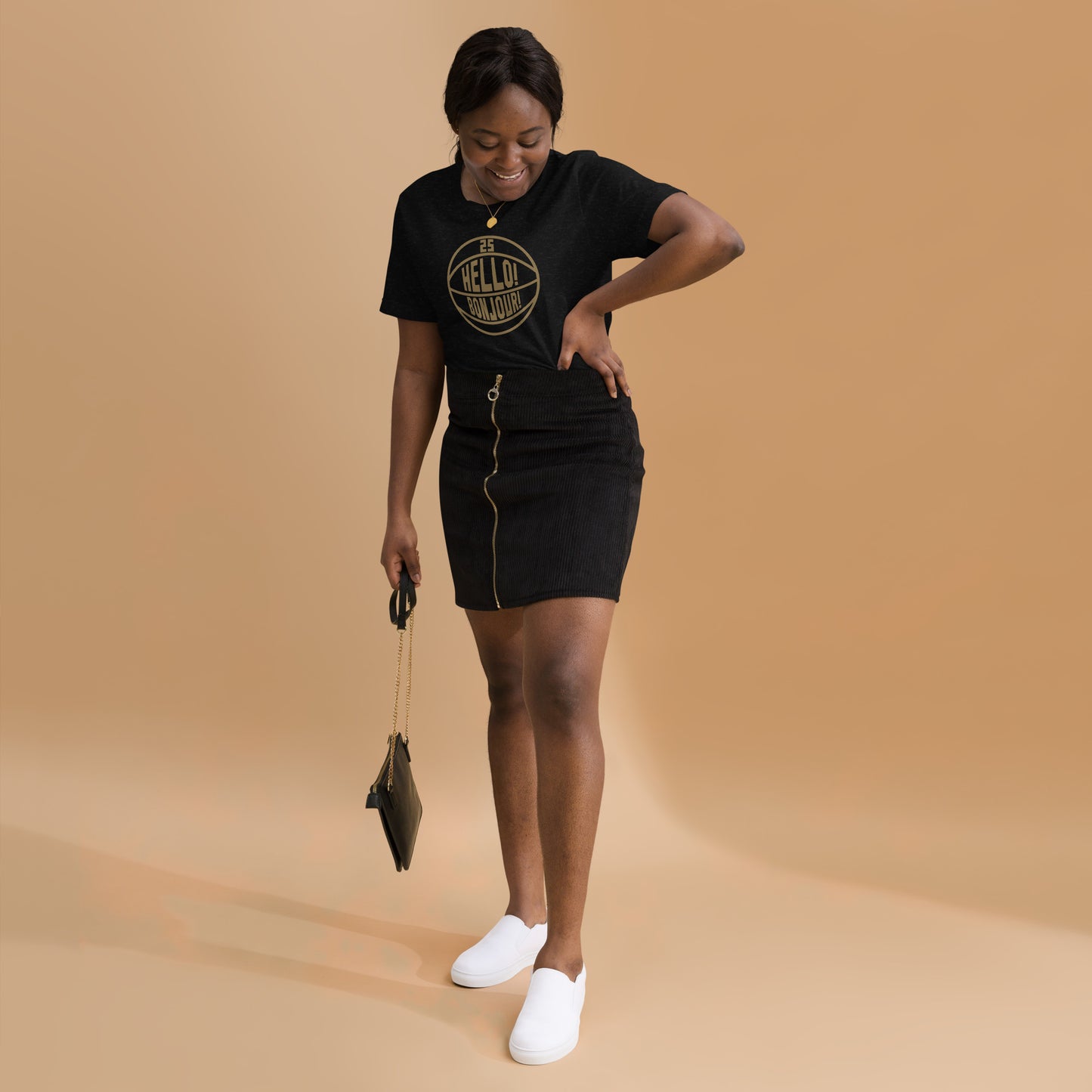 T-shirt worn by young woman, this image details the graphic design "Hello! Bonjour!". In the image, the words are built into a basketball and feature Chris Boucher's number 25. The Toronto Raptors' announcer, Jack Armstrong, loves to shout out Hello! Bonjour! when Chris Boucher does something exciting on the court. The tee is black and the design is in a gold similar to the Toronto Raptors City Edition colours. This item is exclusive to tailgate mercantile and is available only online.