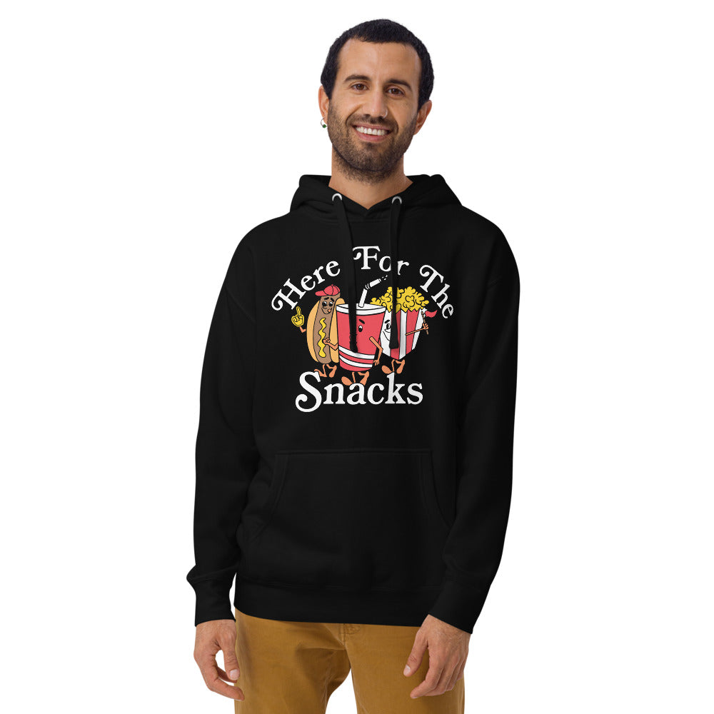 Image of man wearing black hoodie with design of "Here For The Snacks" with cartoon hot dog, soda pop and popcorn located on centre chest. This design is exclusive to Tailgate Mercantile and available only online.