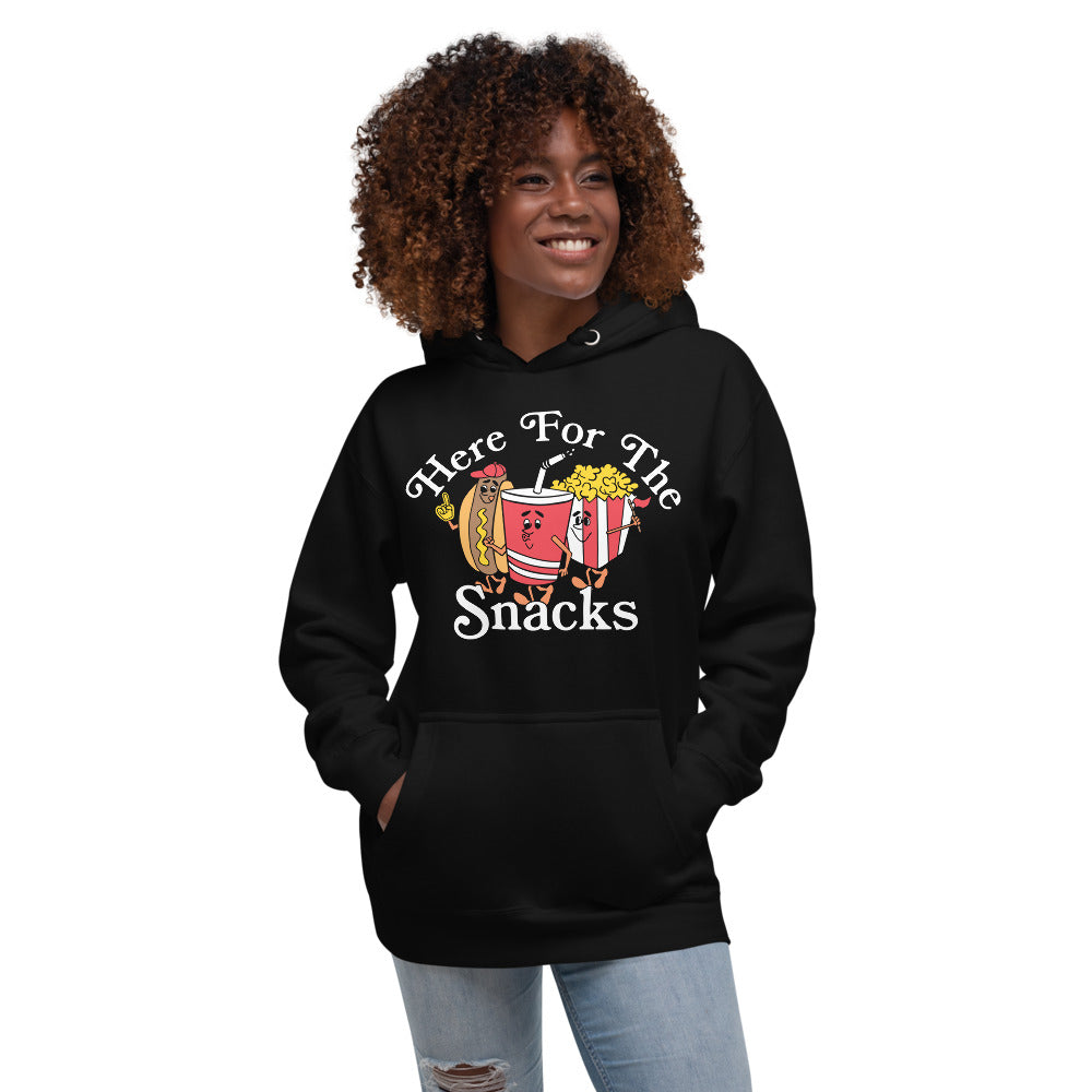 Image of woman wearing black hoodie with design of "Here For The Snacks" with cartoon hot dog, soda pop and popcorn located on centre chest. This design is exclusive to Tailgate Mercantile and available only online.