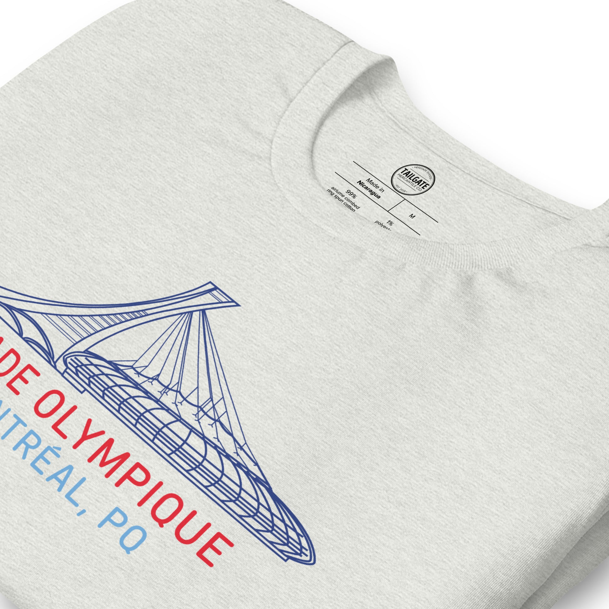Close up image of heather ash t-shirt with design of "Stade Olympique, Montreal, PQ" illustrated ballpark completed in retro Montreal Expos colours located on centre chest. Image includes neck tag information. This design is exclusive to Tailgate Mercantile and available only online.