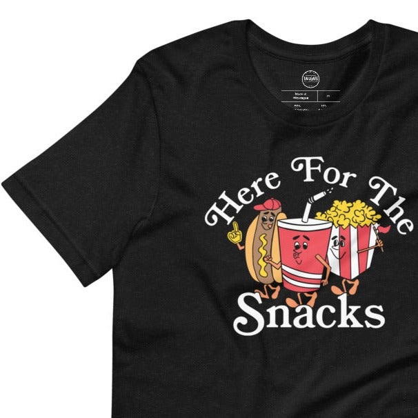 Image of heather black tee with design of "Here For The Snacks" with cartoon hot dog, soda pop and popcorn located on centre chest. This design is exclusive to Tailgate Mercantile and available only online.