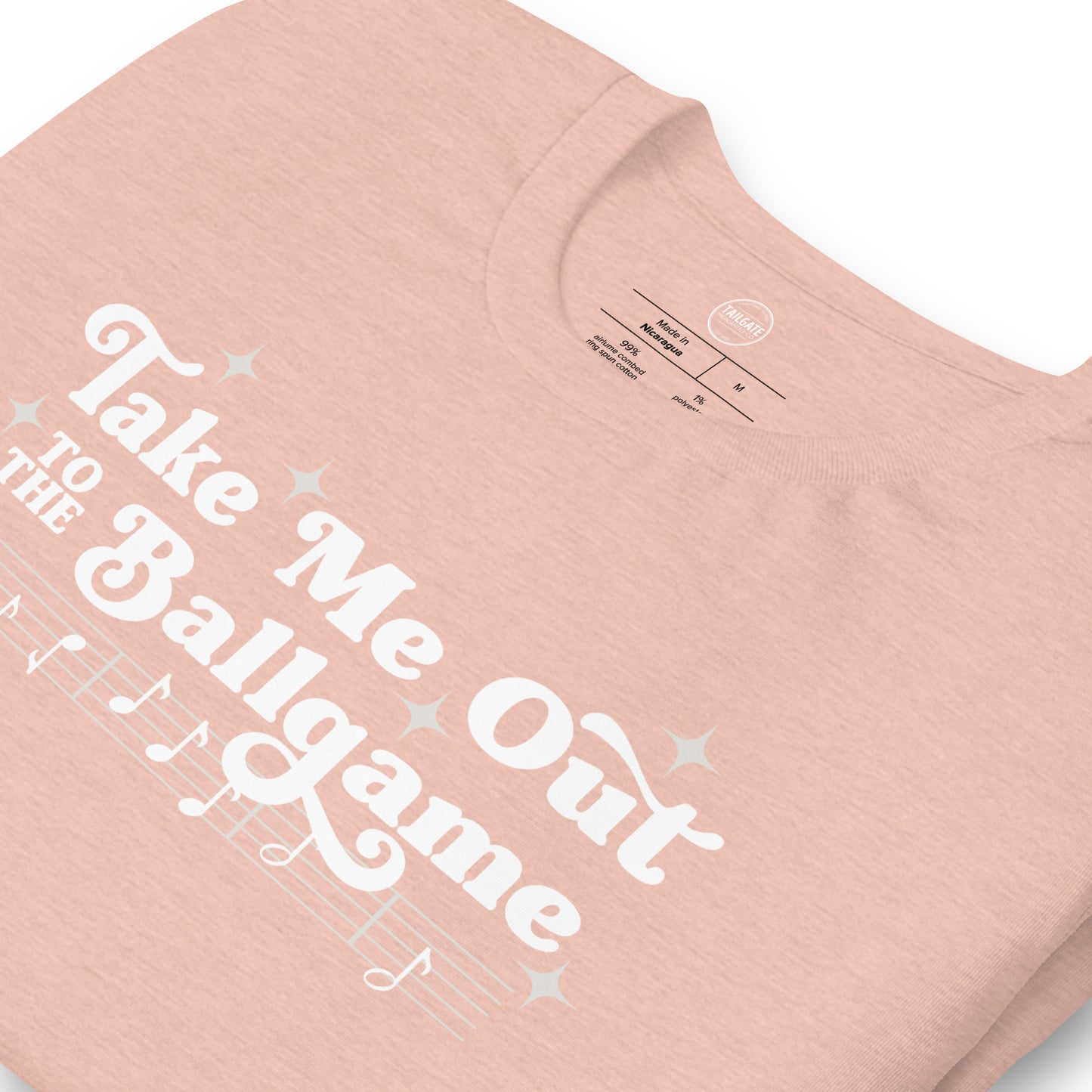 Close up image of heather peach t-shirt with design of "Take Me Out to the Ballgame" with coordinating musical notes in white located on centre chest. This design is exclusive to Tailgate Mercantile and available only online.
