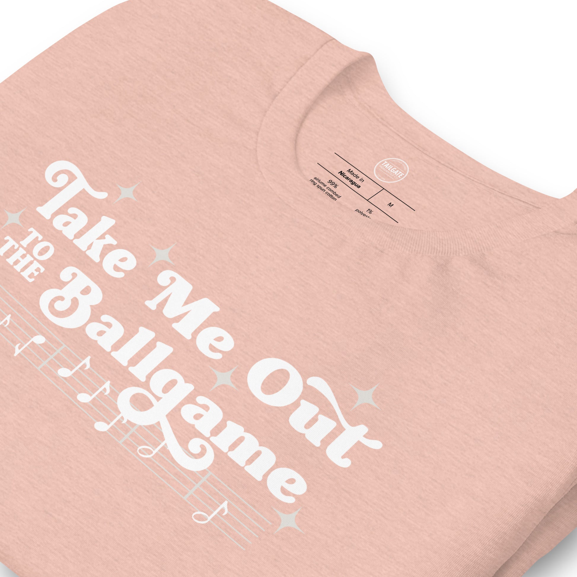 Close up image of heather peach t-shirt with design of "Take Me Out to the Ballgame" with coordinating musical notes in white located on centre chest. This design is exclusive to Tailgate Mercantile and available only online.